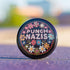 Round pinback metal button featuring an embroidered-style design on a black background. The center showcases bold, stitched-textured lettering in red, blue, and white that reads "PUNCH NAZIS." Surrounding the text is an intricate, folk-inspired floral pattern with multicolored flowers in shades of pink, orange, blue, and white, arranged symmetrically. A decorative zigzag border frames the edge of the button. The backside is silver metal with a secure pin closure.